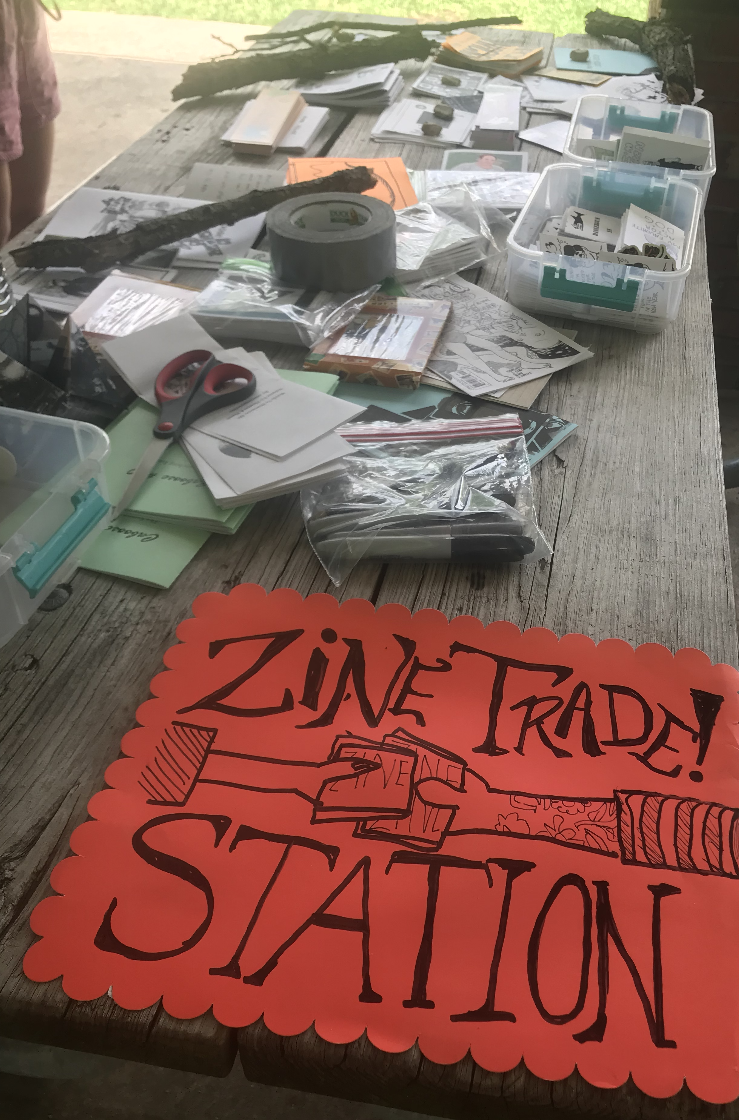 photograph of a table covered in zines with a sign reading "zine trade station"