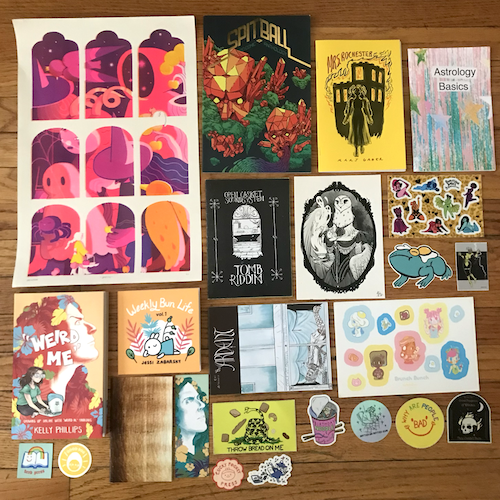 photograph of Alex's CAKE haul on a hardwood floor including mini zines, stickers, and prints