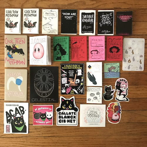 photograph of 1/2 of Alex's CZF haul on a hardwood floor including mini zines, stickers, and prints